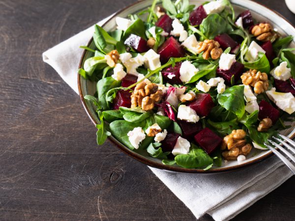 Beet or beetroot salad with fresh arugula, soft cheese and walnuts on plate, dressing and spices on dark wooden background, copy space, top view/